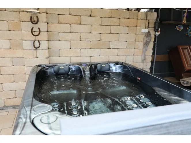 Cadă cu hidromasaj PUMA 210×170×90 cm, cu un design compact, situată într-un mediu interior, având un finisaj negru și dotată cu două șezlonguri ergonomice și un loc de ședere, cu apă în mișcare și un perete de piatră în fundal.