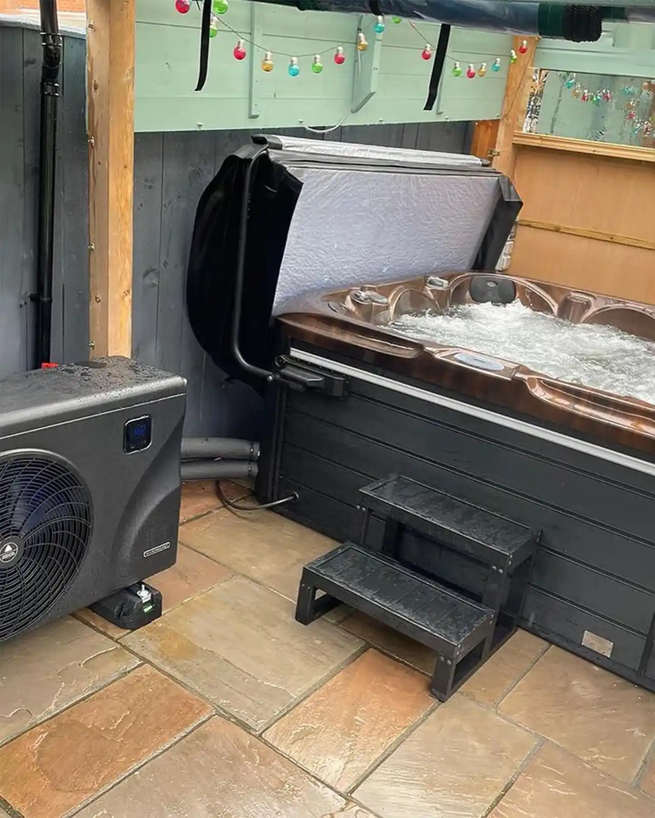 Pompă de căldură ZEALUX pentru jacuzzi, situată lângă un jacuzzi cu apă, pe o podea din piatră, cu un set de trepte negre în față și un panou de protecție deasupra jacuzzi-ului.
