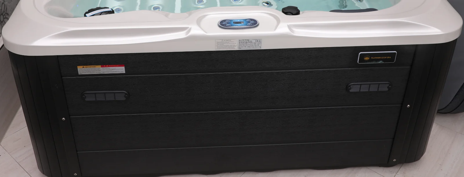 Cadă cu hidromasaj New Maximus DELUXE, cu carcasă neagră și suprafață acrilică albă, având un panou de control digital și un design modern, situată într-un mediu interior cu podea deschisă.