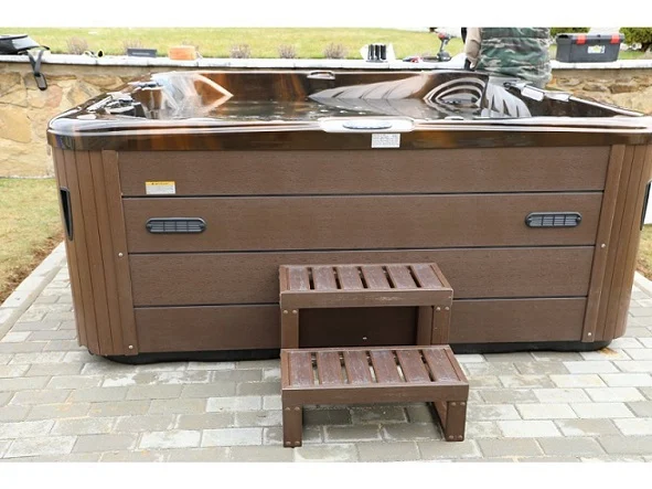 Cadă cu hidromasaj New Maximus, de culoare maro, cu trepte din lemn, situată pe o suprafață de piatră, având un design modern și un aspect elegant, potrivită pentru 4-5 persoane.
