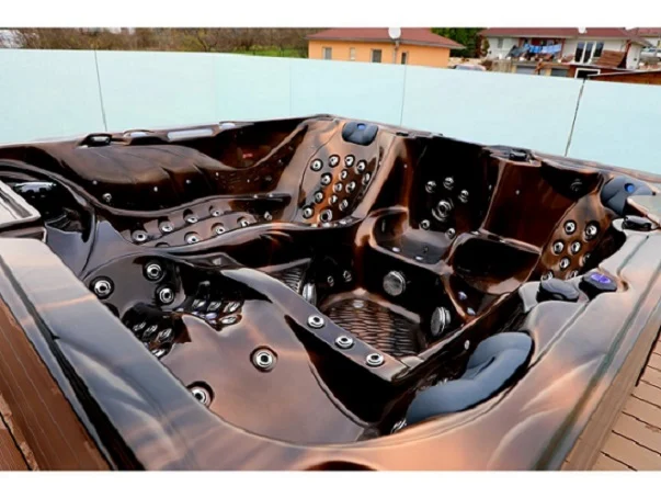 Cadă cu hidromasaj New Maximus, cu un finisaj lucios de culoare cupru, are un design modern și este dotată cu numeroase jeturi de masaj, fiind concepută pentru 4-5 persoane, amplasată pe o terasă din lemn.