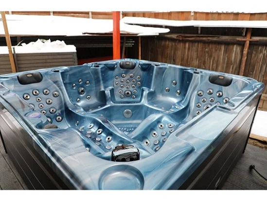 Cadă cu hidromasaj New Maximus, de culoare albastră, cu un design modern, având un interior cu 136 de jeturi de masaj, situată într-un mediu exterior, pe o platformă cu un fundal de lemn și zăpadă.