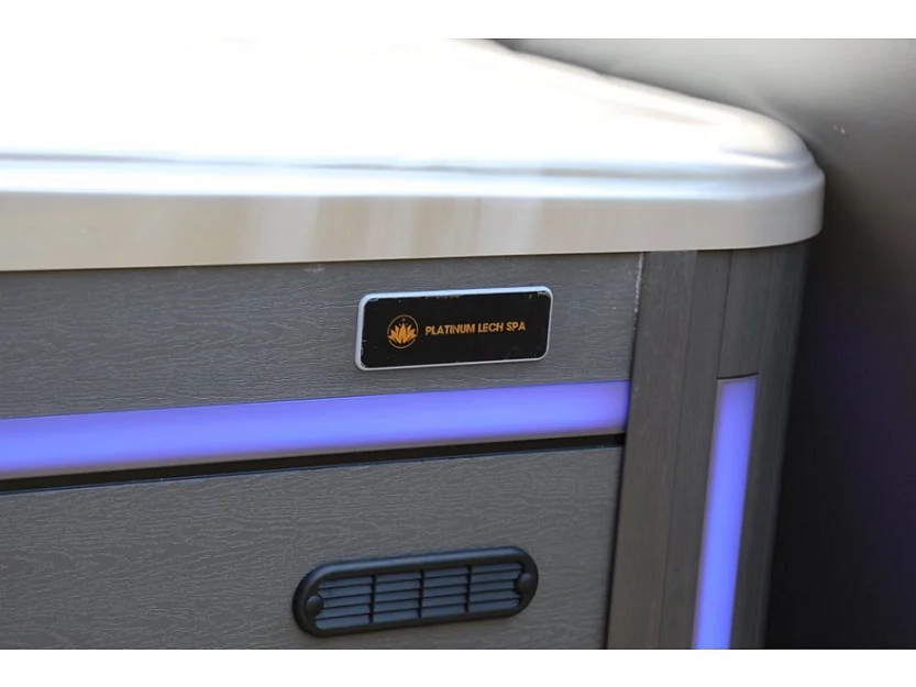 Cadă cu hidromasaj New Maximus cu un finisaj modern, având un panou de control cu iluminare albastră și un logo elegant, situată într-un mediu luminos, cu un design din acril de culoare gri.