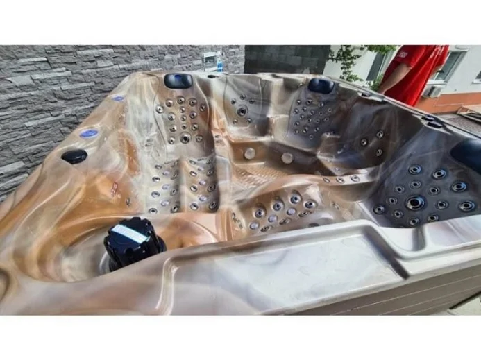 Cadă cu hidromasaj New Maximus, cu un design modern, din acril de culoare deschisă, având 136 de jeturi de masaj, situată într-un mediu exterior cu pereți din piatră.