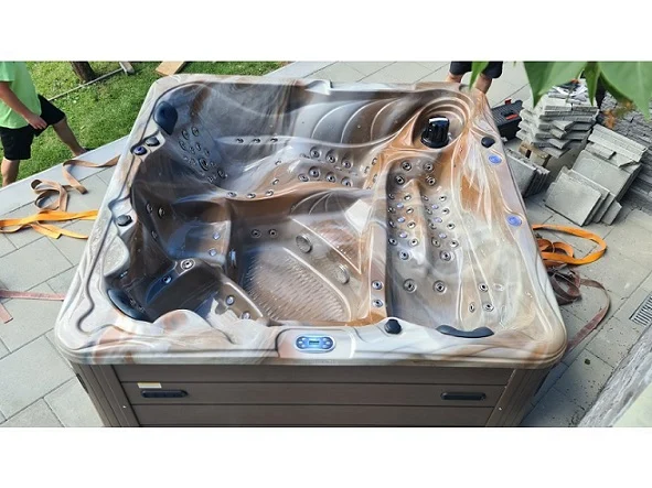 Cadă cu hidromasaj New Maximus, cu un design modern, de culoare maronie cu nuanțe deschise, având 136 de jeturi de masaj, amplasată pe o platformă de piatră, înconjurată de iarbă și materiale de construcție.