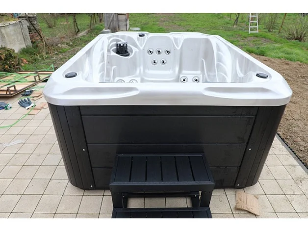 Cadă cu hidromasaj SUN 200×160×90 cm, cu un design compact, de culoare albă și neagră, situată pe o platformă de piatră, având 3 locuri, inclusiv 2 șezlonguri și 1 loc de ședere, înconjurată de un peisaj verde.