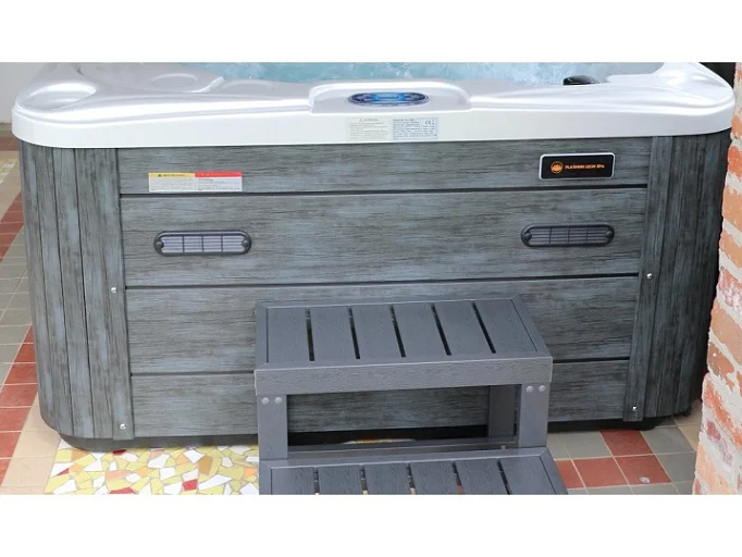 Cadă cu hidromasaj SUN 200×160×90 cm, cu carcasă gri și trepte din lemn, amplasată pe o podea cu mozaic colorat, având un panou de control pe margine.