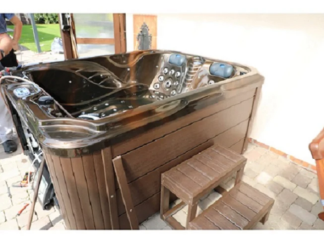 Cadă cu hidromasaj SUN 200×160×90 cm, cu un design compact, din acril negru, având 3 locuri, este amplasată pe o terasă, lângă o ușă de intrare, și este însoțită de două trepte din lemn.