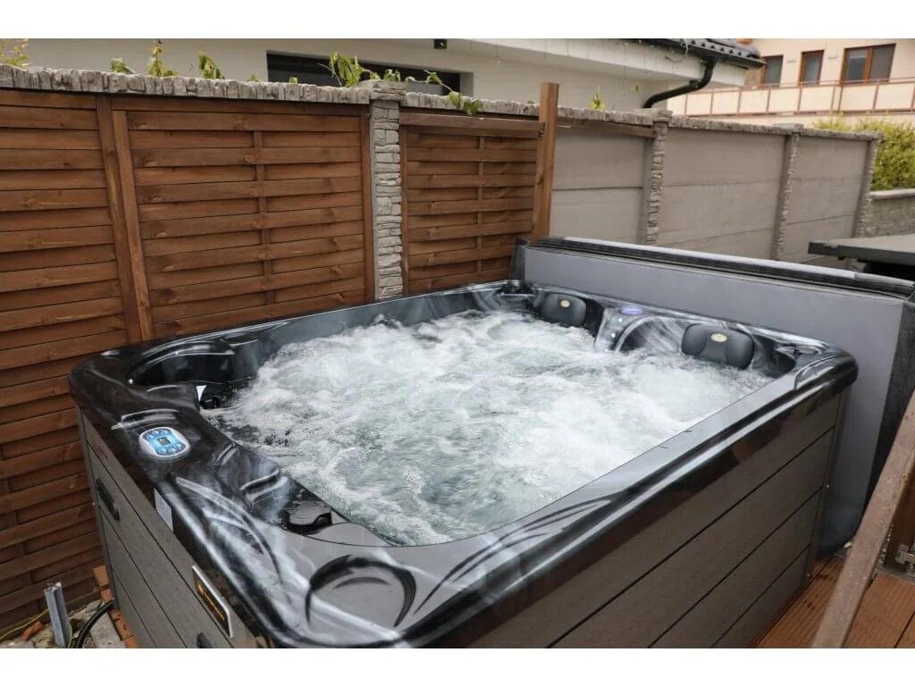 Cadă cu hidromasaj PUMA 210×170×90 cm, cu un design compact, este vizibilă într-un mediu exterior, având un finisaj negru lucios și un interior cu apă în mișcare, fiind înconjurată de un gard din lemn și un perete de piatră.