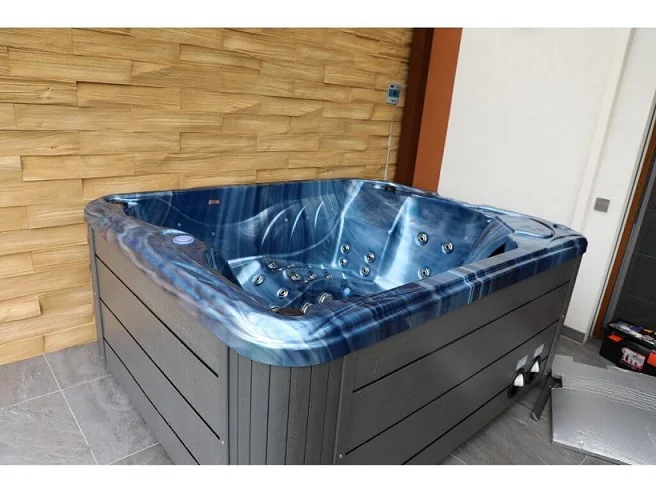 Cadă cu hidromasaj PUMA 210×170×90 cm, cu un design compact, are un interior de culoare albastră cu un model de valuri, fiind amplasată pe o podea din gresie gri, într-un mediu modern cu pereți din lemn.