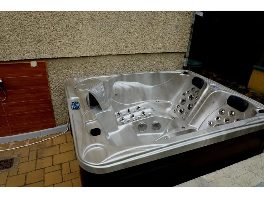 Cadă cu hidromasaj PUMA 210×170×90 cm, cu un design compact, din material acrilic, având un interior argintiu cu duze de masaj, situată pe o terasă cu pavaj de piatră, înconjurată de un perete texturat.