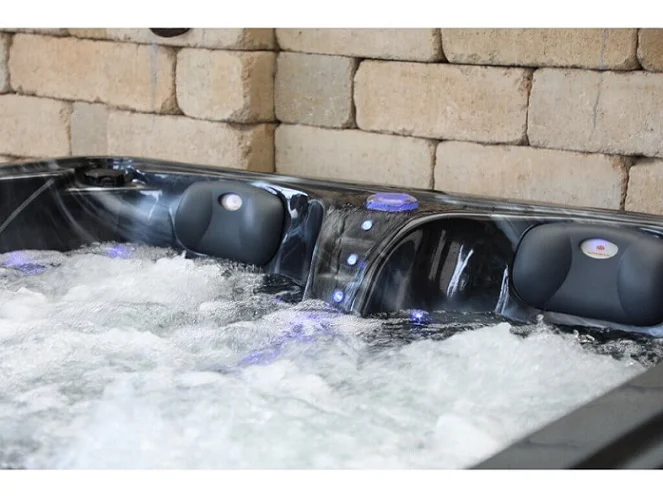 Cadă cu hidromasaj PUMA 210×170×90 cm, cu un design compact, are două șezlonguri ergonomice și un loc de ședere, fiind evidențiată de un finisaj negru lucios și lumini LED în jurul marginilor, amplasată într-un mediu cu pereți din piatră.