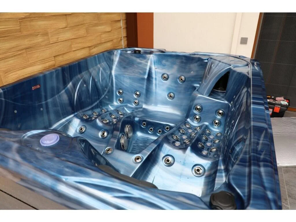 Cadă cu hidromasaj PUMA 210×170×90 cm, de culoare albastră cu un model fluid, dotată cu numeroase jeturi de masaj, situată într-un mediu interior cu pereți din lemn și un aspect modern.