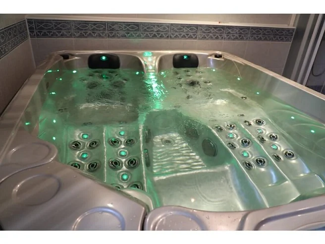 Cadă cu hidromasaj PUMA 210×170×90 cm, cu un interior din acril transparent, iluminată cu lumini verzi, având două șezlonguri și un loc de ședere, situată într-un mediu interior cu plăci ceramice.