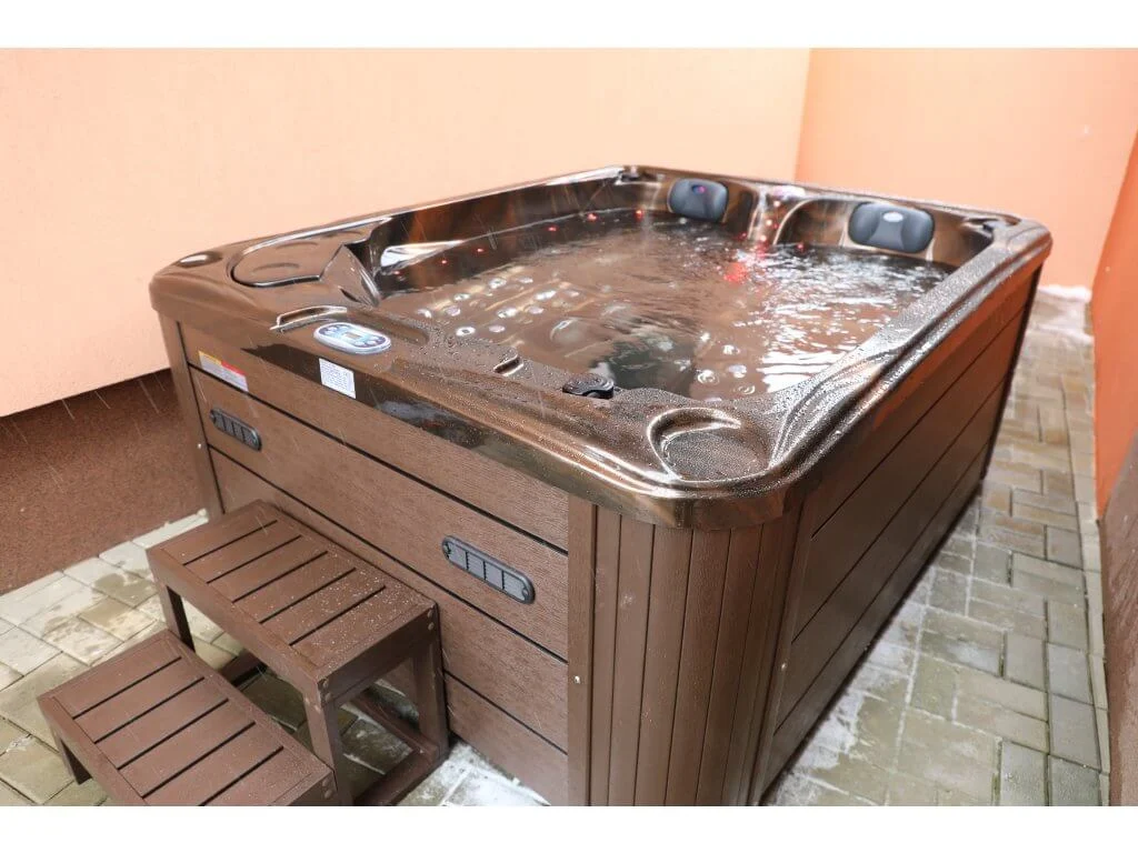 Cadă cu hidromasaj PUMA 210×170×90 cm, de culoare maro, cu un design compact, situată pe o terasă, având două șezlonguri ergonomice și un loc de ședere, cu trepte din lemn pentru acces.