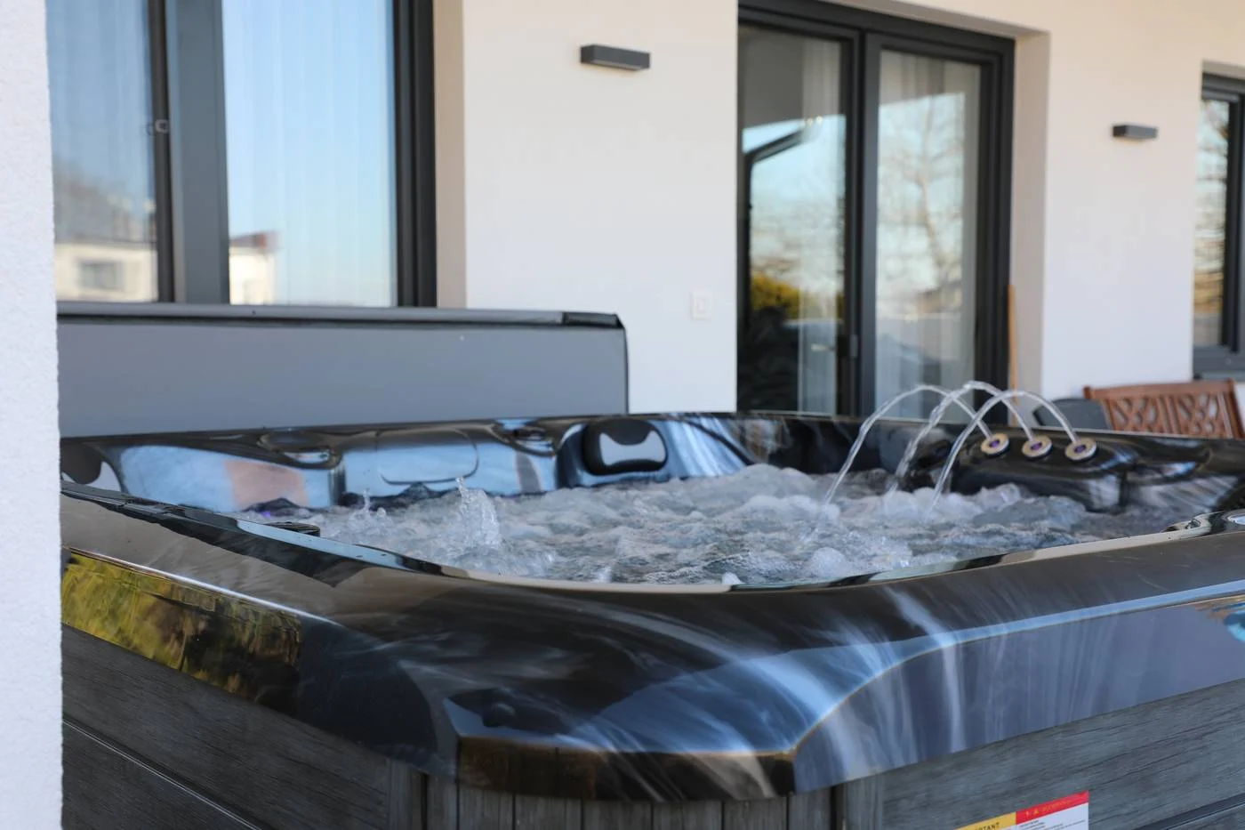 Cadă cu hidromasaj LIFE 220×220×90 cm, cu un design elegant, de culoare neagră, situată pe o terasă, cu apă în mișcare și jeturi de masaj vizibile, înconjurată de feronerie modernă și feronerie de sticlă.