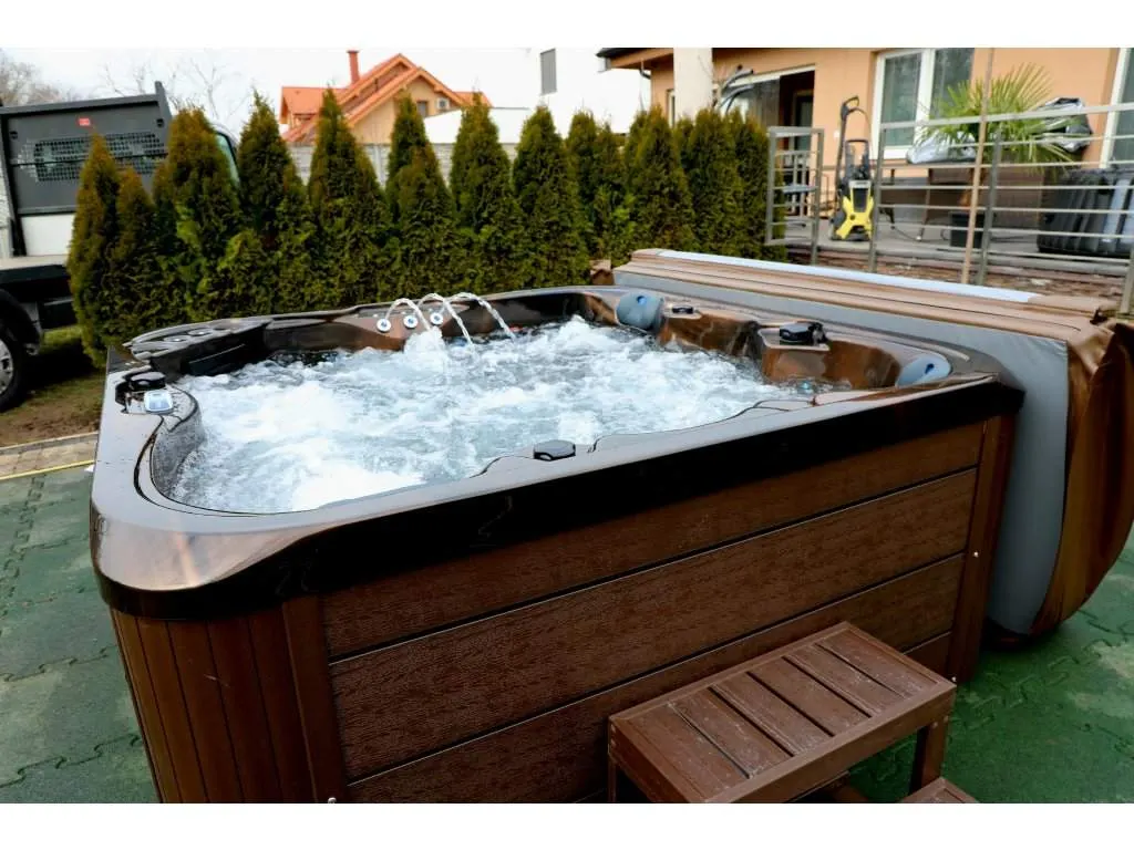 Cadă cu hidromasaj SPLIT 200×200×90 cm, cu un design modern și ergonomic, situată într-o grădină, având un exterior din lemn de culoare maro și un interior cu apă în mișcare, fiind dotată cu jeturi de masaj, ideală pentru 4-5 persoane.