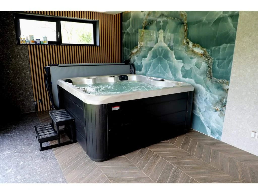 Cadă cu hidromasaj SPLIT 200×200×90 cm, cu carcasă neagră, amplasată într-o încăpere modernă, având un perete decorat cu un model de piatră verde, lângă o fereastră mare, și cu scări din lemn pentru acces.