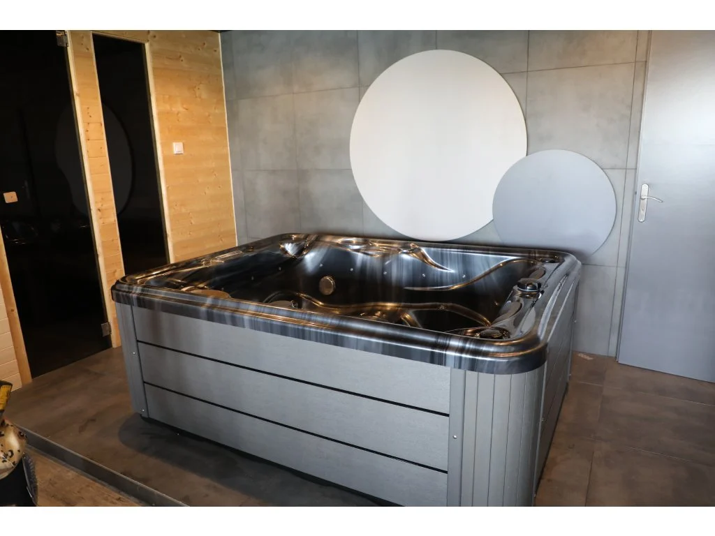 Cadă cu hidromasaj compactă HOME 210×160×77 cm, cu un design modern, situată într-un spațiu interior, având un finisaj gri și un cadru din material acrilic, ideală pentru 2-3 persoane.